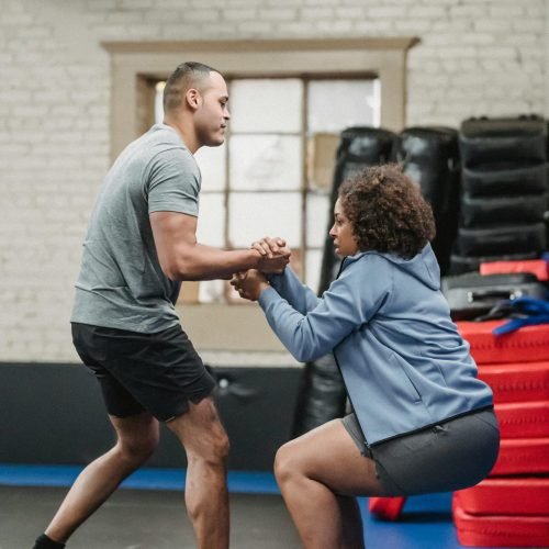 A personal trainer helps a client up in a fitness studio, surrounded by workout equipment.