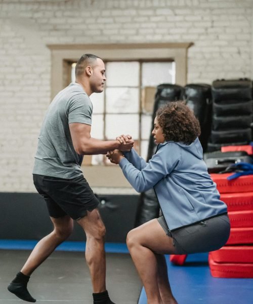 A personal trainer helps a client up in a fitness studio, surrounded by workout equipment.
